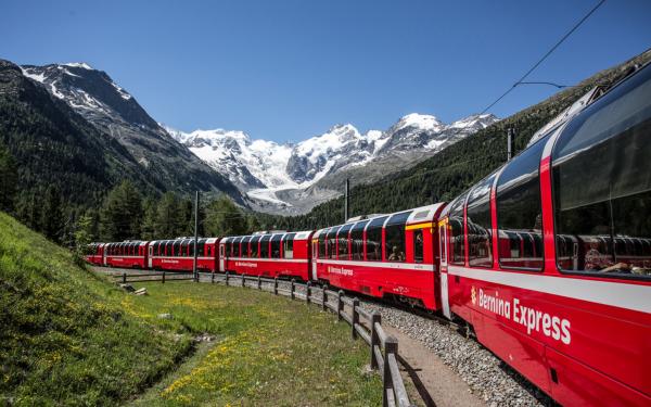 The Bernina Express train travelling through a spring landscape with snowy mountains behind